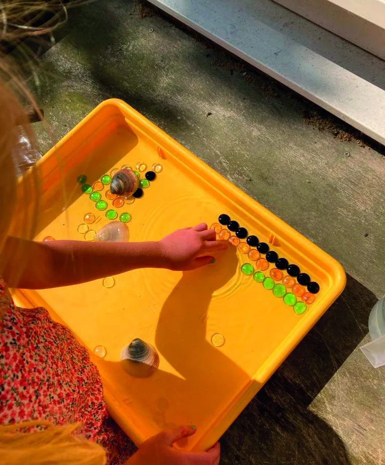 Kind speelt met water, knikkers en schelpen in een gele waterbak